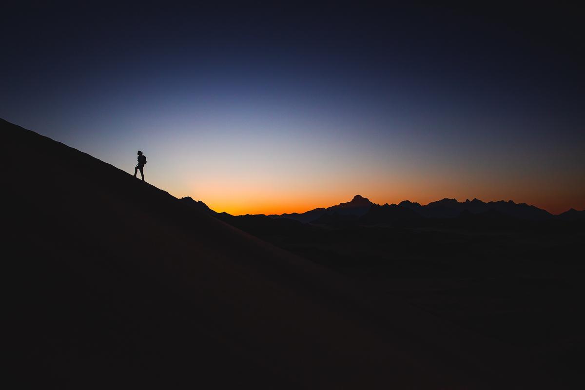 Illustration of a person with a backpack standing on top of a mountain, looking at the sunset over a beautiful landscape.